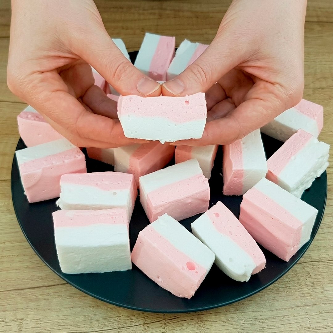 Hands holding pink and white striped marshmallow bites cut into neat blocks arranged on a dark plate